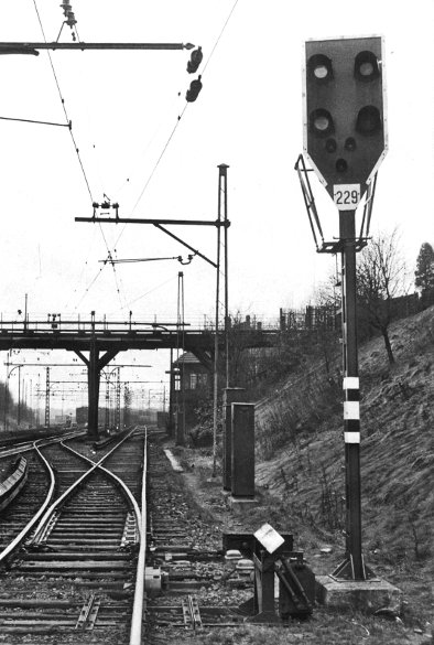 Graustufenfoto des Sv-Signals 229 mit vier Laternen rechts vom S-Bahngleis nach Ohlsdorf vor einer Weichenspitze, darüber die Oberleitung, rechts eine ansteigende Böschung, im Hintergrund eine Straßenbrücke über die Gleise